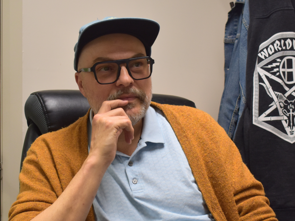 Chris Friesen, PARC's Manager of Collaborative Care, on mental health support in Parkdale. He is wearing a baseball cap, thick rimmed glasses, a yellow cardigan, and light blue button-down shirt.