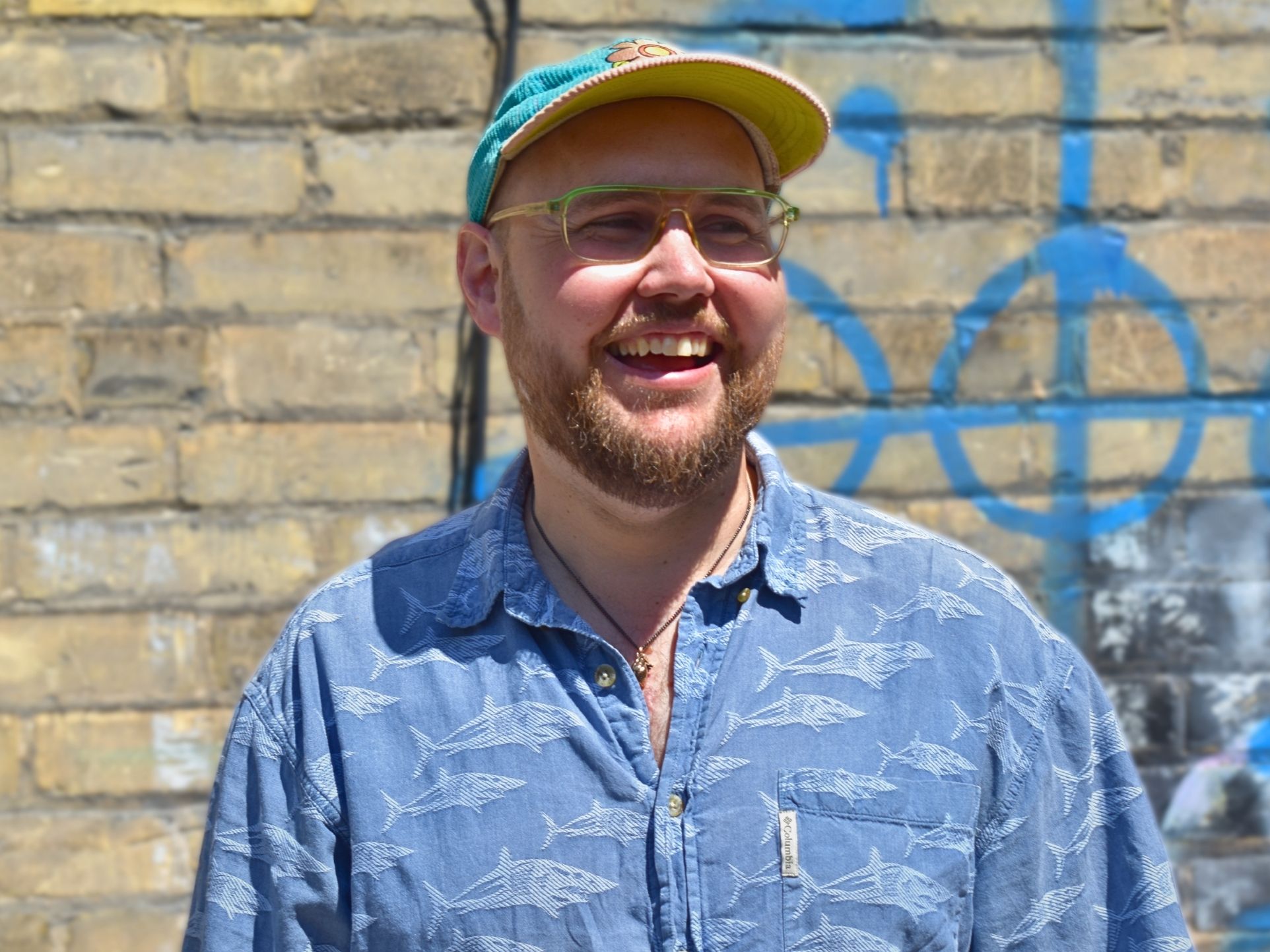 A white man in glasses is smiling at something off camera. He is wearing a blue and yellow baseball cap, and a blue collared shirt with a shark pattern.
