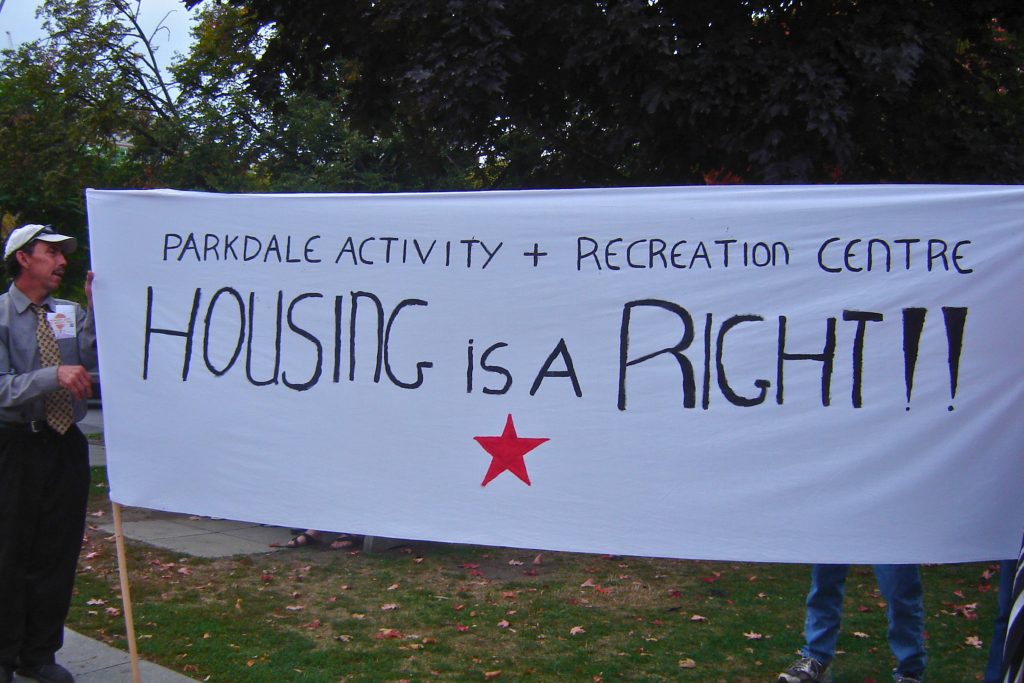 A sign is being held up in protest of the affordable housing crisis in Toronto. A white banner reads "Parkdale Activity-Recreation Centre" and "HOUSING is a RIGHT!!" with a red star on the banner.